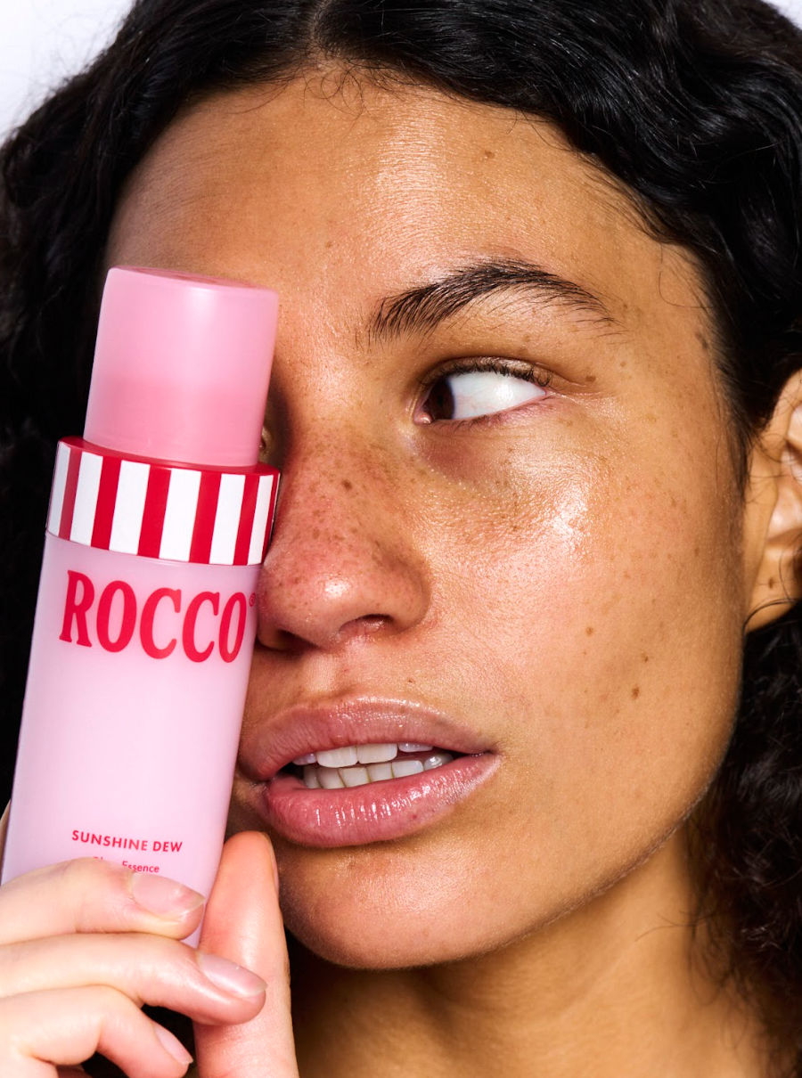 Person holding a pink and white striped bottle labeled 'Rocco' near their face.