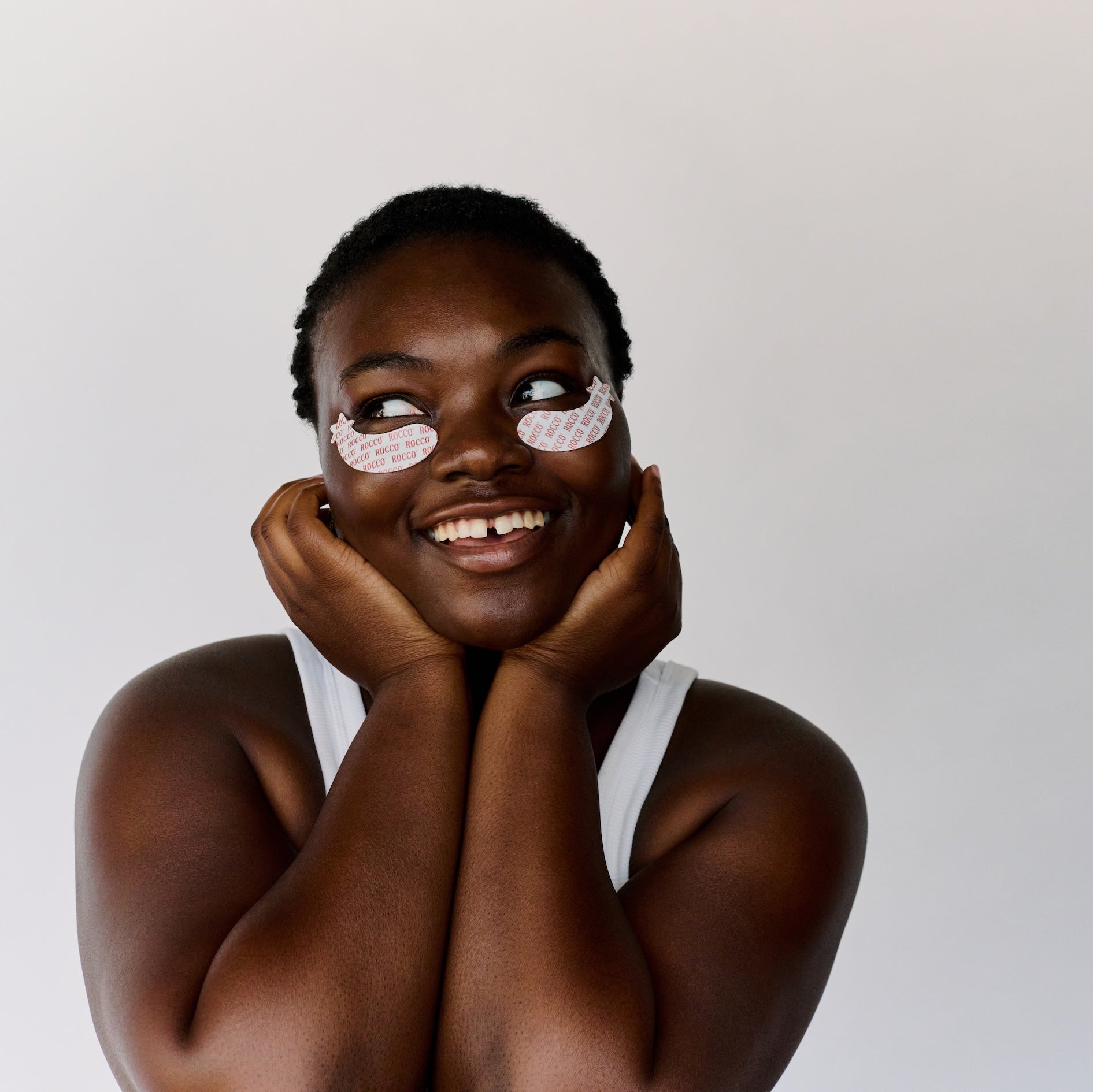 A smiling woman with ROCCO Happy Eyes  eye patches under her eyes poses against a plain light gray background.