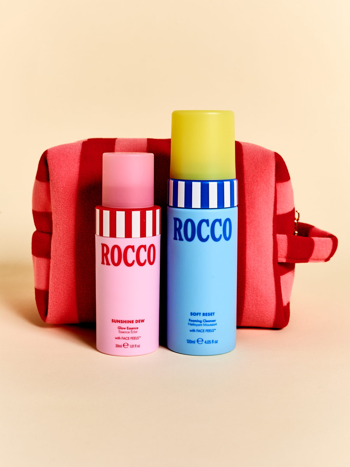 Two Rocco fragrance bottles standing in front of a red-and-white striped toiletry bag on a warm beige studio background. The bottle on the left is pink with red-and-white stripes and the word ROCCO, and the bottle on the right is light blue with blue-and-white stripes and the words ROCCO and SOFT RESET.