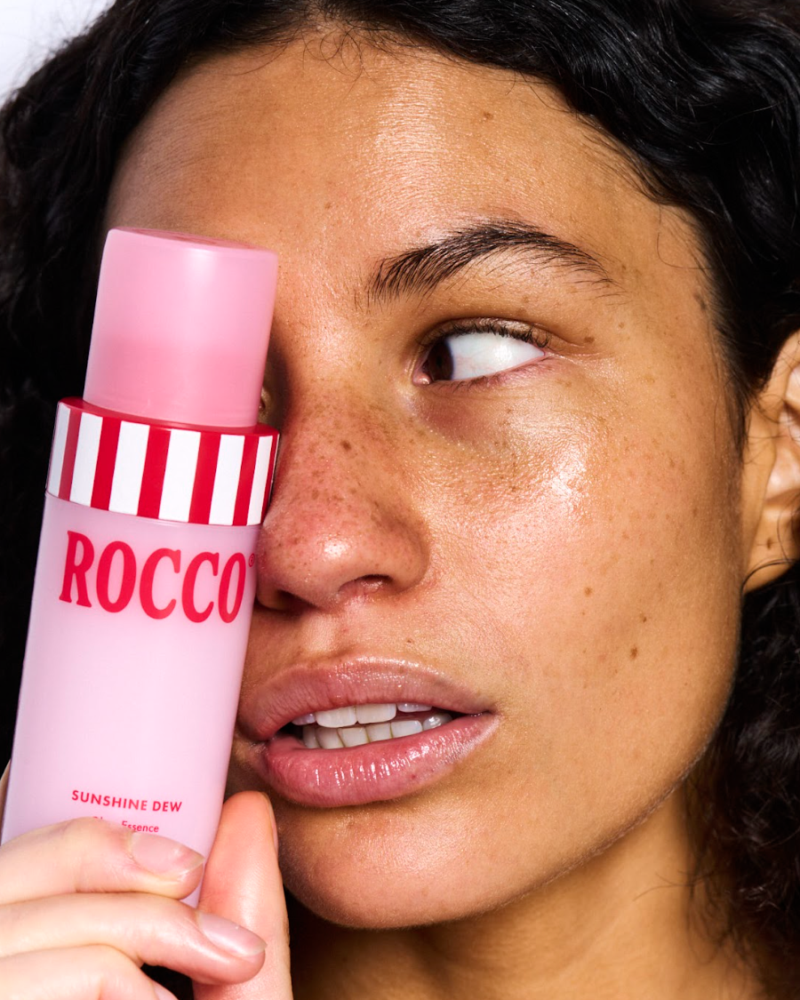 Person holding a pink and white striped bottle labeled 'ROCCO Sunshine Dew' near their face.